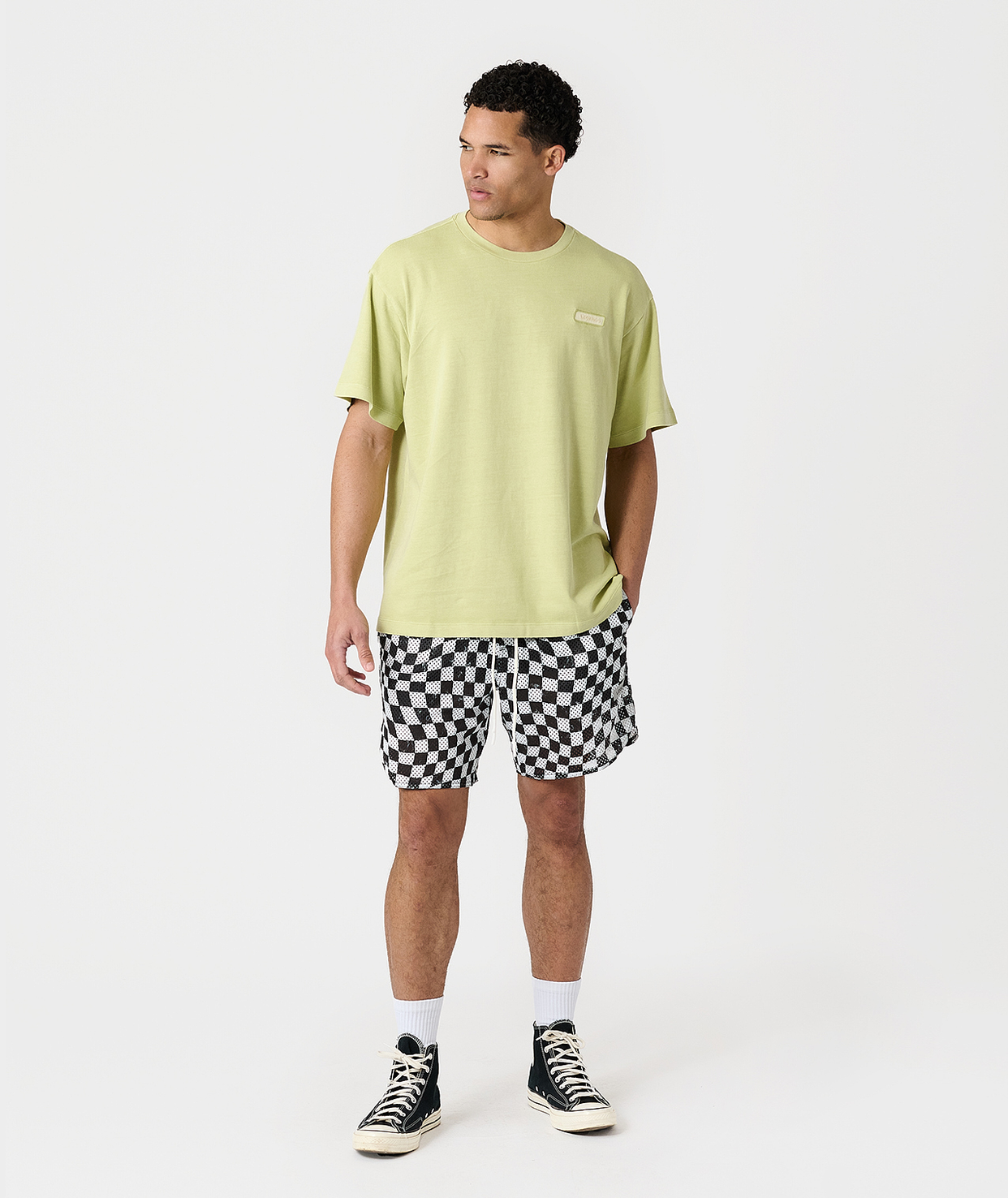 Man wearing lime green t-shirt and black-and-white checkerboard mesh shorts with white socks and black canvas sneakers.