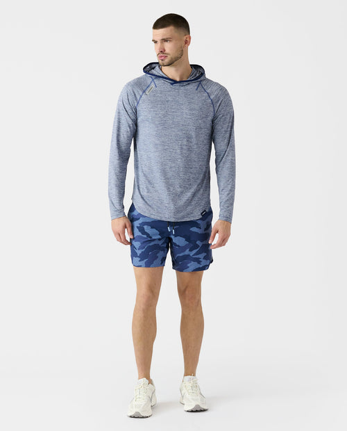 Man wearing ocean heather hooded long-sleeve top with blue camouflage shorts and white sneakers.